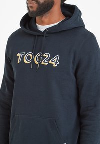Man wearing navy blue hoodie with front pocket and "TOG24" logo in white and yellow on chest, white shirt visible underneath.