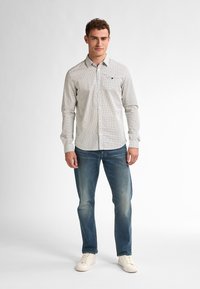 Camicia bianca con pois neri, maniche lunghe e una piccola tasca sul petto, abbinata a jeans azzurri chiari e sneaker bianche.