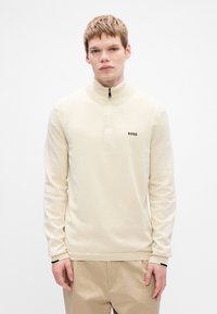 Cream half-zip sweatshirt with black logo on chest, ribbed cuffs, and hem; smooth texture, fitted design. Model wearing light trousers.