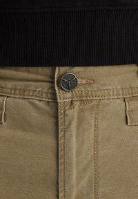 Beige jeans feature a textured fabric with a black ribbed waistband, metal button closure, and standard pocket design with visible stitching.