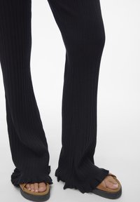 Pantalons noirs côtelés avec un ourlet évasé, en tissu texturé. Associés à des sandales à glissière tan, laissant apparaître les orteils.