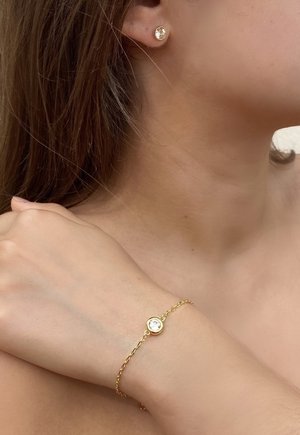 Goldfarbene Ohrringe und ein Armband mit kreisförmigen Designs, die einen klaren, funkelnden Stein in der Mitte haben. Detail des Kettenarmbands sichtbar.
