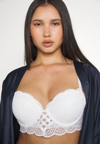 Soutien-gorge en dentelle blanche présentant un motif géométrique, un soutien-armature et des bords festonnés ; bretelles ajustables et un détail transparent au centre.