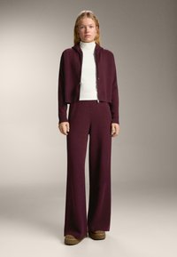 Žena stojí oblečená v bielej roláku, burgundskej mikine na zips, širokých burgundských nohaviciach a hnedých platformových topánkach na jednoduchom pozadí.