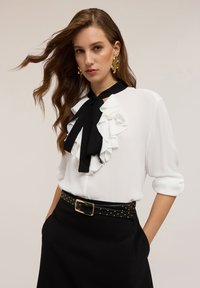 Blusa bianca con accenti arricciati neri e cravatta, abbinata a una gonna nera con borchie dorate e cintura con fibbia. Texture liscia.
