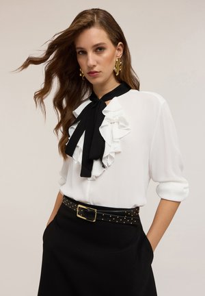 Blusa bianca con accenti arricciati neri e cravatta, abbinata a una gonna nera con borchie dorate e cintura con fibbia. Texture liscia.