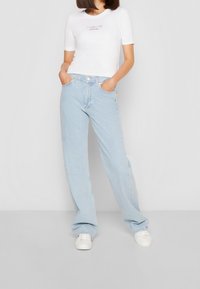 Žena v světle modrých širokých džínách, bílých teniskách a bílém tričku s krátkým rukávem s nápisem "Toto je láska Calvin Klein", s rukama v kapsách.