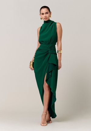 Vrouw draagt een mouwloze jurk in smaragdgroen met hoge split en gouden accessoires, staand tegen een eenvoudige lichte achtergrond.
