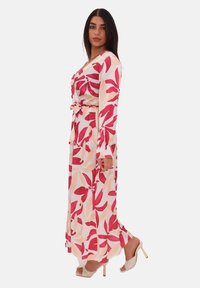 Robe longue à motifs floraux avec des feuilles roses et pêches, fabriquée en tissu léger. Dotée d'une taille ceinturée et de manches longues. Portée avec des sandales à talons.