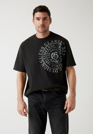 Un tricou negru din bumbac cu guler rotund și mâneci scurte, având un grafic circular mare de culoare gri pe față, împreună cu text. Este purtat cu blugi închiși la culoare.