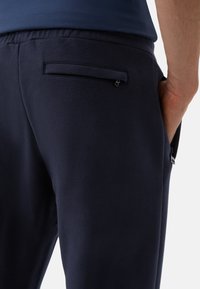 Tmavě modré teplákové kalhoty z měkké látky, s kapsou na zadní straně se klopou a bočními kapsami pro praktické využití a pohodlí.