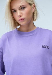 Lila Baumwoll-Sweatshirt mit Rundhalsausschnitt und kurzen Ärmeln, mit "1980" in Schwarz auf der Brust bedruckt. Glatte Textur.