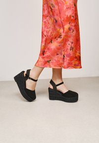 Sandalias de cuña negras con suela de yute texturizado, correa ajustable en el tobillo y parte superior de tela suave. Llevadas con un vestido floral rosa.