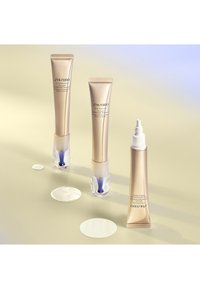 Tre Shiseido Vital Perfection-kräm-tuber i guldfärgade tuber med genomskinliga bottnar, omgivna av krämklickar på en ljus bakgrund.