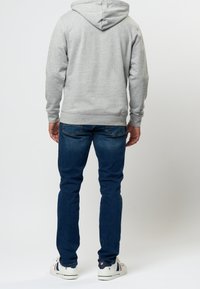 Felpa grigia con cappuccio dotata di coulisse e tasca a marsupio, abbinata a jeans blu scuro e sneakers bianche con dettagli blu navy e rossi.