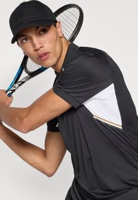 Μαύρο αθλητικό πόλο με κοντά μανίκια, λευκές και χρυσές λεπτομέρειες, υφασμάτινη υφή και εφαρμοστή σχεδίαση. Ένα άτομο κρατά μια ρακέτα τένις σε θέση έτοιμη για παιχνίδι.