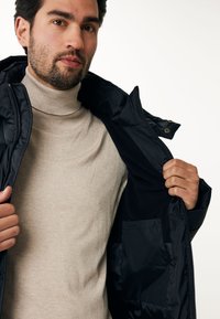 Chaqueta de plumas negra con capucha, cierre frontal de cremallera y interior suave en contraste. Cuello de tortuga de color beige claro debajo. Se puede ver un bolsillo vertical.