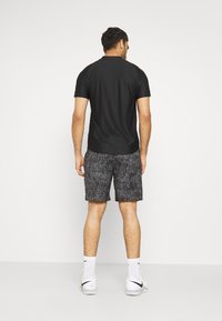 Schwarzes kurzärmeliges T-Shirt und gemusterte schwarze Shorts. Glatter Stoff, lockere Passform, kombiniert mit weißen Sportschuhen und weißen Socken in Knöchellänge.