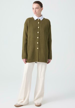 Ženska, oblečena v olivno zeleno rebrasto jopico z gumbi, belo srajco z ovratnikom, kremne široke hlače in bež čevlje, stoji na belem ozadju.