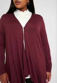 Femme portant des boucles d'oreilles en or, une blouse blanche avec un col noué, et un cardigan long ouvert bordeaux.