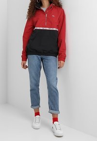 Osoba v červené a černé polozipové bundě, s vyhrnutými modrými džíny, červenými ponožkami a bílými teniskami stojí proti bílé zdi.