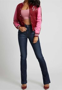 Bordeaux satijnen varsityjas met roze mouwen, witte letter "G" detail; gecombineerd met donkerblauwe flare jeans en rode schoenen.