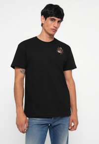 T-shirt en coton noir avec un graphisme de spider coloré sur la poitrine gauche. Manches courtes et design classique, associé à un jean en denim.