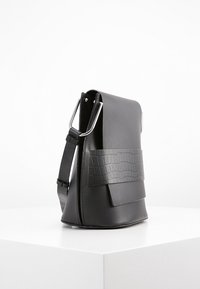 Černá kožená taška s texturovaným krokodýlím vzorem. Má elegantní obdélníkový tvar a stříbrné kovové připevnění popruhu.