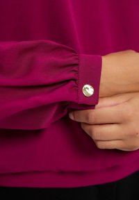 Fuksjowa bluzka z długimi, bufkami, ze zebranymi mankietami i złotym akcentem w postaci guzika. Gładka tkanina o delikatnym połysku i wyraźnie ustrukturyzowanym kroju.