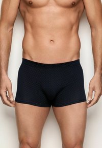 Torso masculino y parte superior de las piernas vistiendo bóxer oscuros de color azul marino con pequeños puntos rojos, de pie contra un fondo claro y liso.