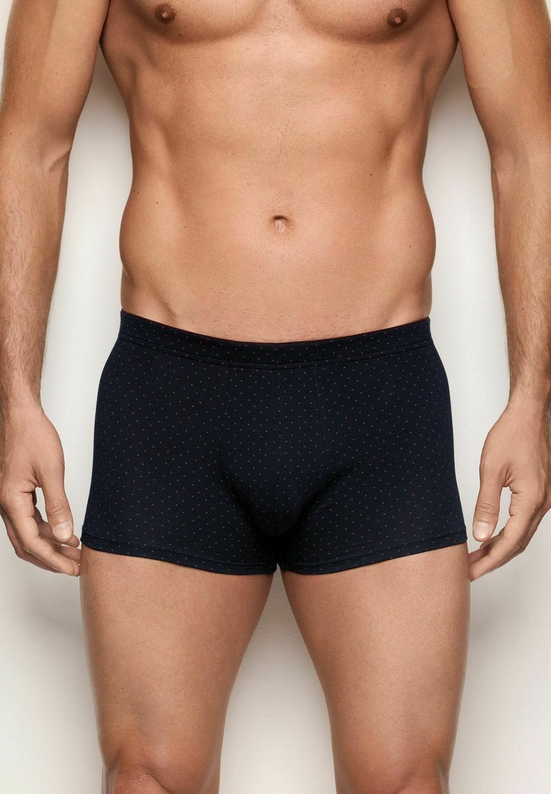 Torso masculino y parte superior de las piernas vistiendo bóxer oscuros de color azul marino con pequeños puntos rojos, de pie contra un fondo claro y liso.