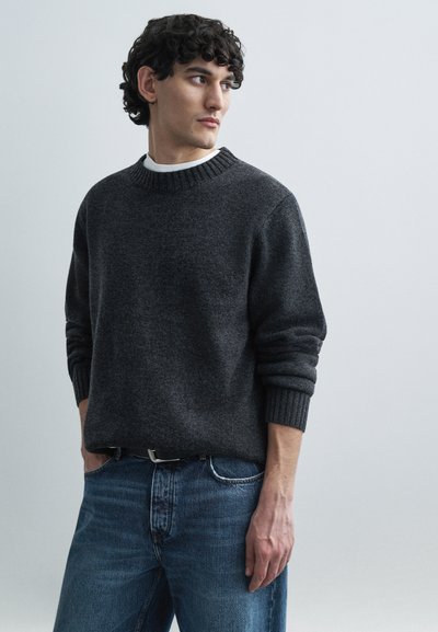 Pull en maille grise avec un col et des poignets côtelés, associé à un jean en denim bleu. Le pull a une coupe décontractée et un tissu texturé.