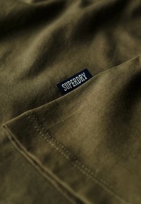 Tejido verde olivo con una textura suave, con una etiqueta azul que dice "SUPERDRY" cosida en la esquina. Vista en primer plano.