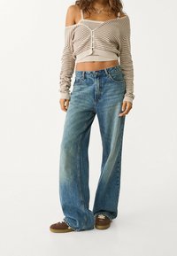 Mujer vistiendo unos vaqueros anchos azules, zapatillas marrones, y un cárdigan corto a rayas beige sin hombros con botones y un collar.