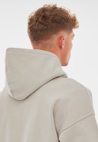 Hanorac gri deschis cu glugă, având o glugă mare din material moale, umeri căzuți și un material neted, ușor texturat.