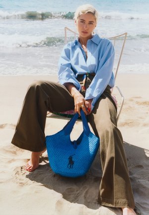 Γυναίκα με κοντά ξανθά μαλλιά κάθεται ξυπόλητη σε καρέκλα σε αμμώδη παραλία, φοράει μπλε πουκάμισο και καφέ παντελόνια, κρατώντας μια μπλε πλεκτή τσάντα.