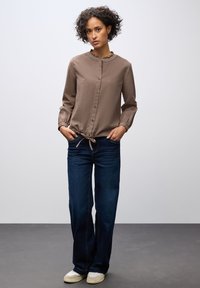 Camicia marrone con bottoni, colletto arricciato e maniche lunghe, abbinata a jeans blu scuro e scarpe chiare. Design semplice e casual.