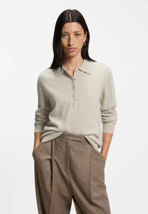 Mujer con cabello largo y oscuro, que lleva un suéter beige abotonado y pantalones marrones holgados con las manos en los bolsillos, posando sobre un fondo blanco.