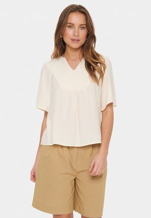Short-sleeve, light beige top with a V-neck and gathered detail. Worn with tan shorts, featuring a relaxed fit and soft texture.