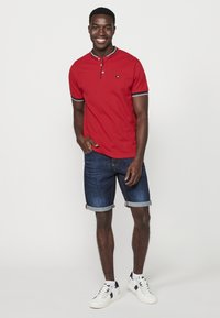 Polo roșu cu mânecă scurtă, cu guler, având un mic ecuson, combinat cu pantaloni scurți din blugi închis la culoare cu manșete și adidași albi.
