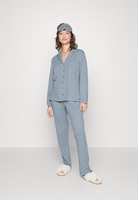 Ljust blå pyjamasset med vita prickar, som inkluderar en skjorta med knappar och byxor med elastisk midja. Mjukt material och matchande ögonmask.