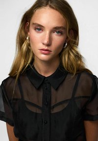 Chemise noire transparente à boutons avec un col classique, des manches courtes et des détails en panneaux courbés subtils au niveau de la poitrine.