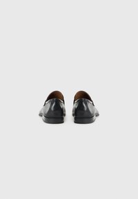 Mocasines de cuero negro con una textura suave, punta redondeada y suela baja y plana. Presentan un mínimo de costuras y un forro interior de cuero marrón.