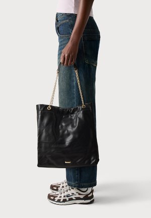 Black quilted leather tote bag with chain straps, featuring a gathered top edge and a gold logo accent at the bottom. Worn with casual jeans and sneakers.