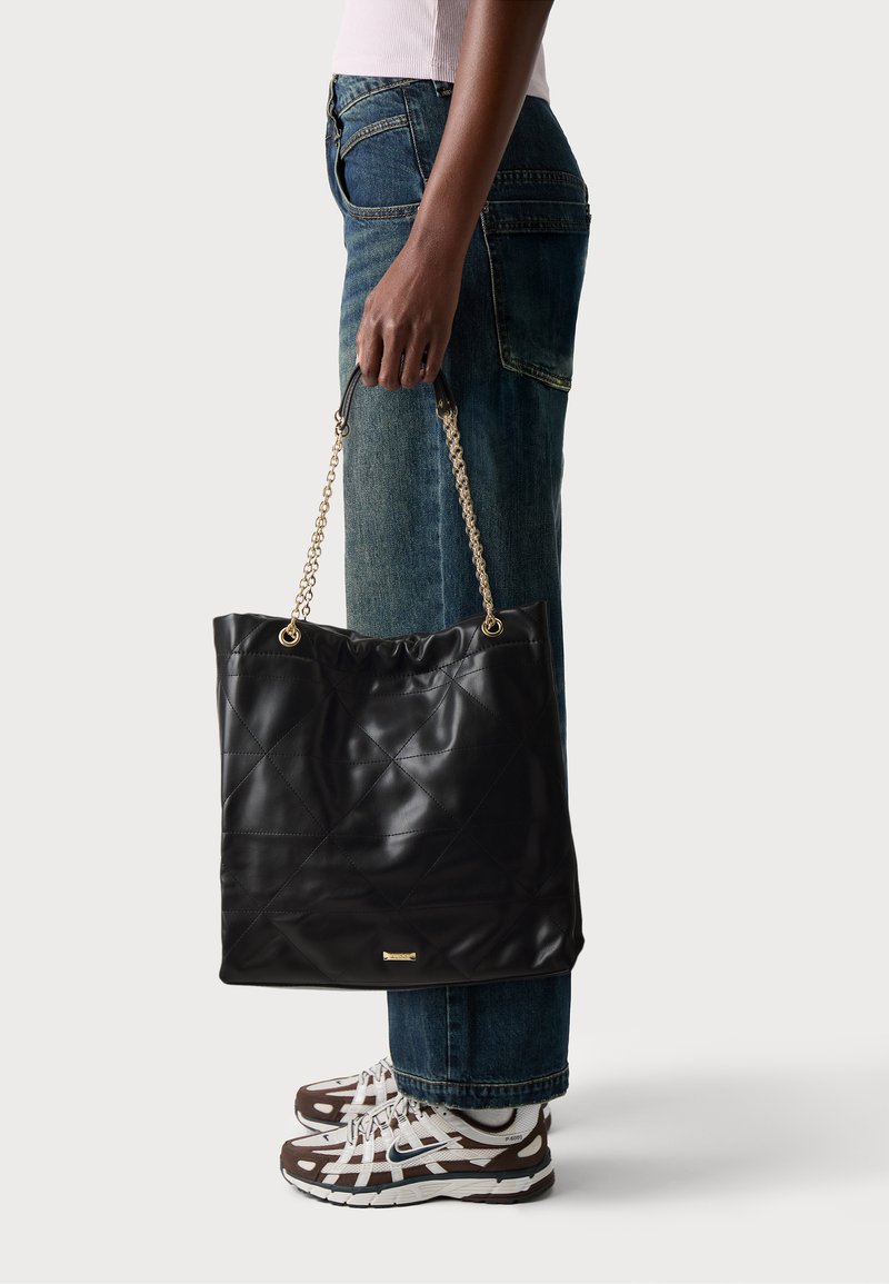 Sac fourre-tout en cuir matelassé noir avec des poignées en chaîne, arborant un bord supérieur froncé et un accent logo doré en bas. Porté avec un jean décontracté et des baskets.