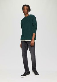 Grüner Cordpullover, runder Ausschnitt, strukturierter Stoff, über ein helles Hemd getragen, kombiniert mit dunkelgrauen Slim-Fit-Jeans und schwarzen Sneakers.