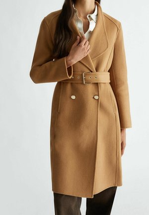 Femme portant un manteau en laine couleur camel ceinturé, par-dessus une chemise blanche boutonnée et un pantalon foncé, sur un fond blanc uni.