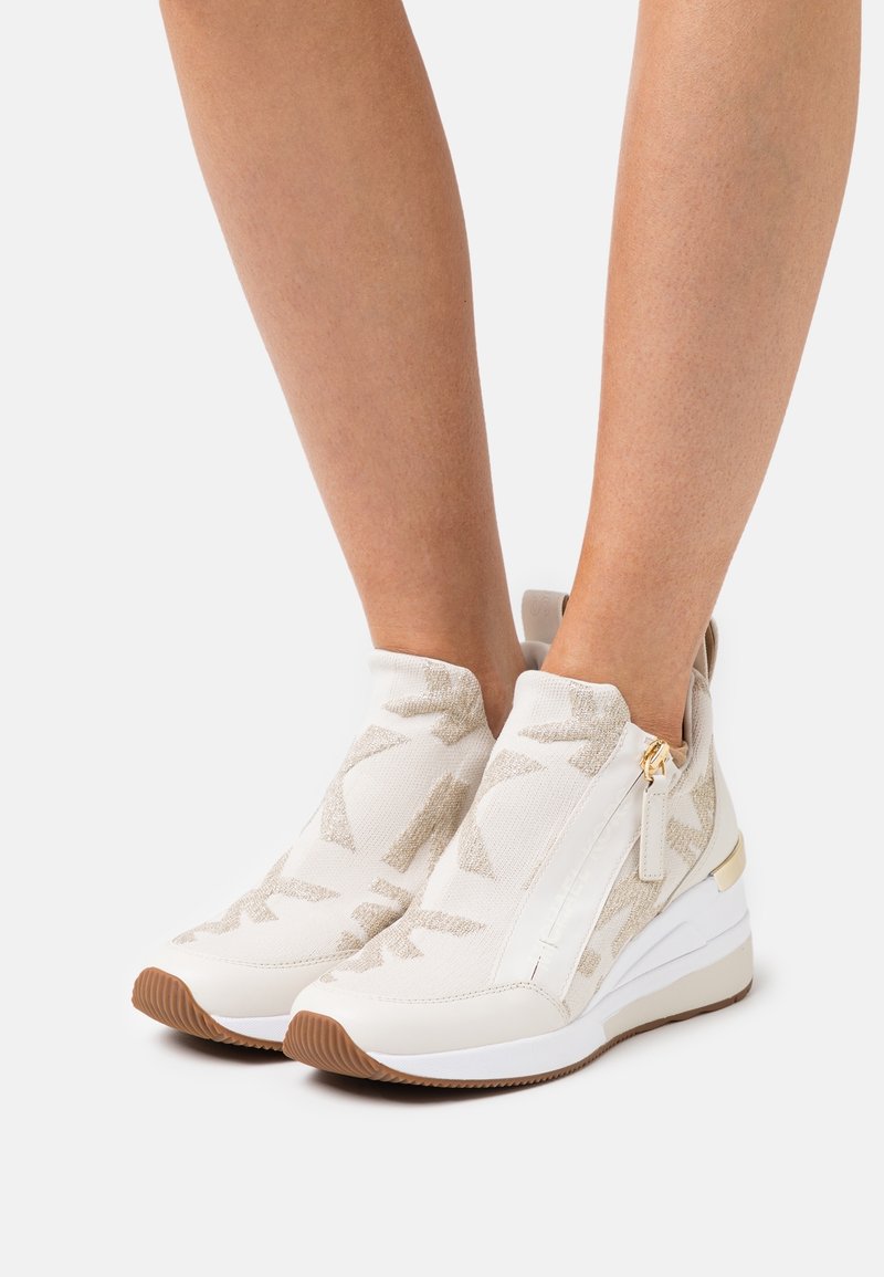 Pieds portant des baskets compensées montantes beige et blanc avec fermetures éclairs latérales et semelles en caoutchouc texturées.
