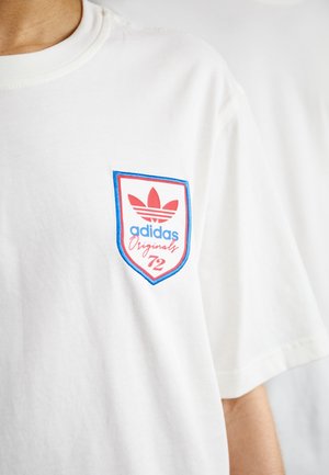 Biała koszulka z niebiesko-czerwonym naszywką z logo Adidas Originals 72 na lewej piersi, noszona przez osobę o jasnej karnacji.