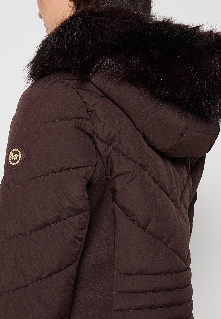 Vrouw in een donkerbruin gewatteerd jack met een bontkraag op de capuchon, zichtbaar MK-logo op de linker mouw.